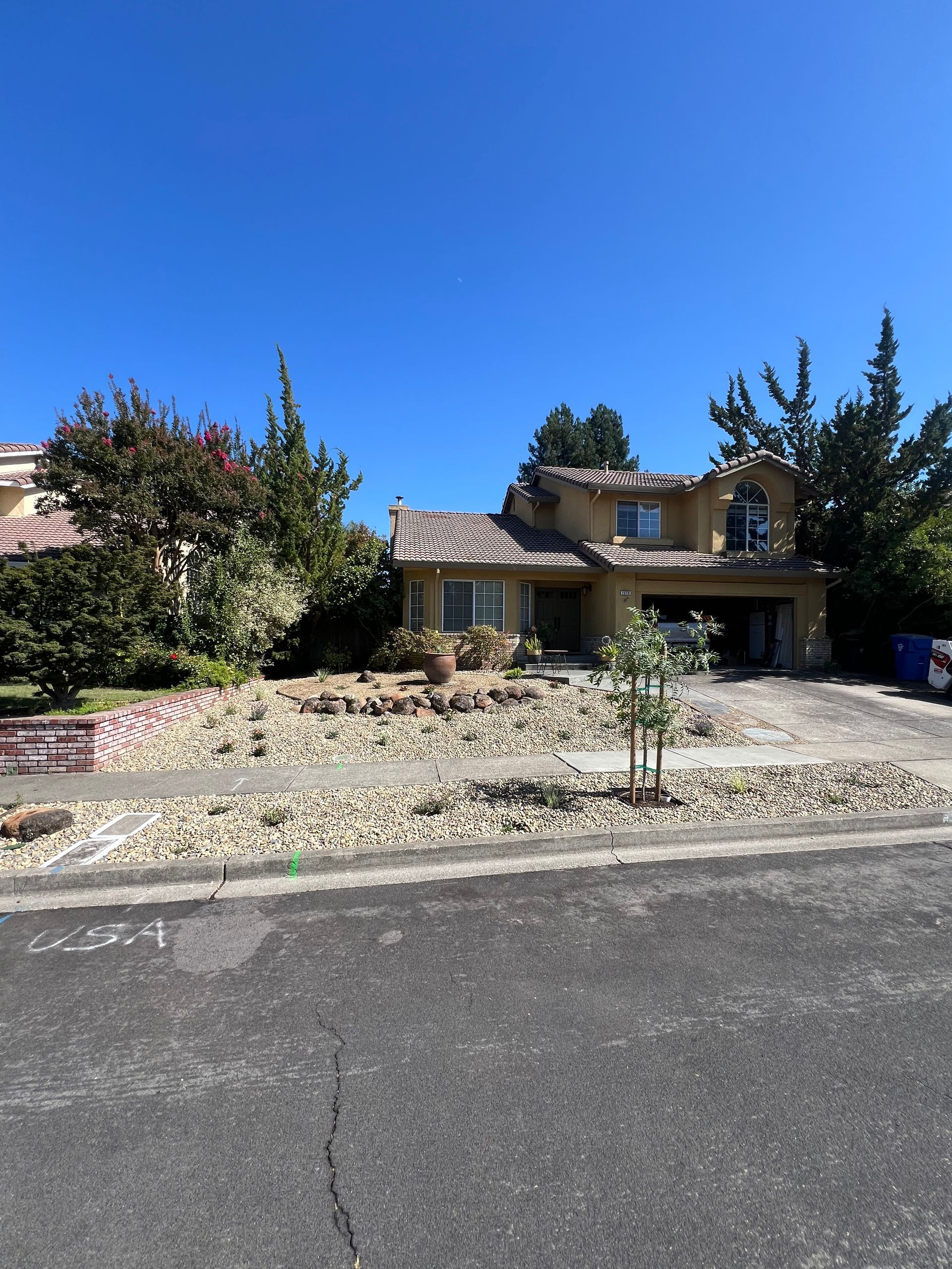 House with damaged roof, debris in yard, and gravel landscaping under a blue sky.