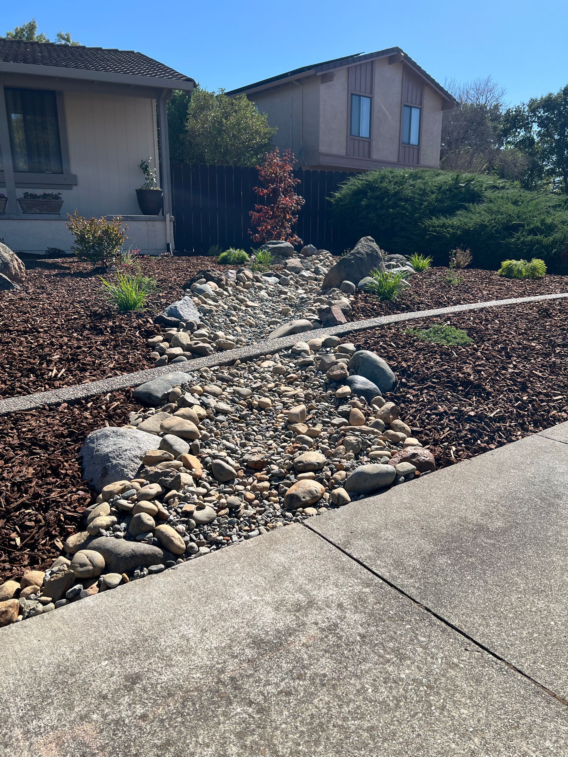 Dry creek bed with rocks and mulch in front yard of homes on a sunny day.