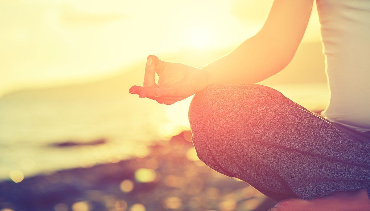Person meditating with hands in a mudra pose, outdoors at sunset.