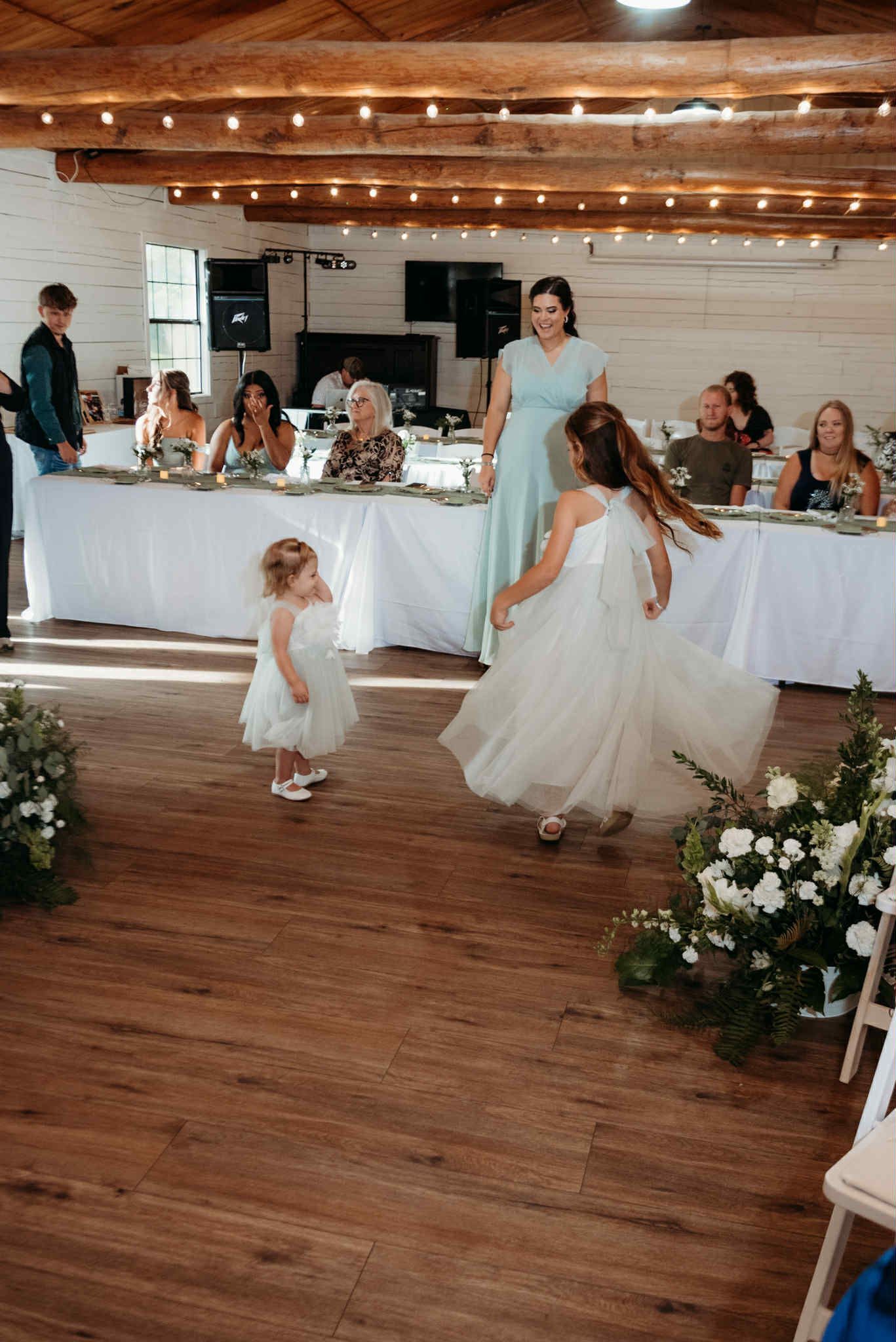 Two flower girls spin on a wooden dance floor during a wedding reception, in front of a long table of guests.