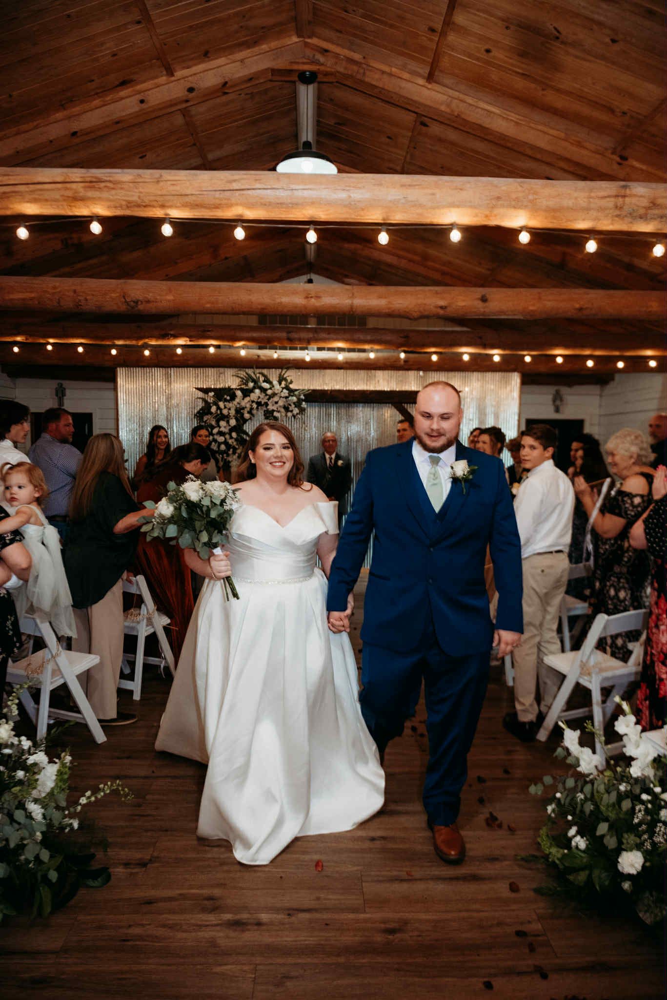 Newlyweds walk down aisle, holding hands. Bride in white dress, groom in blue suit. Ceremony setting.