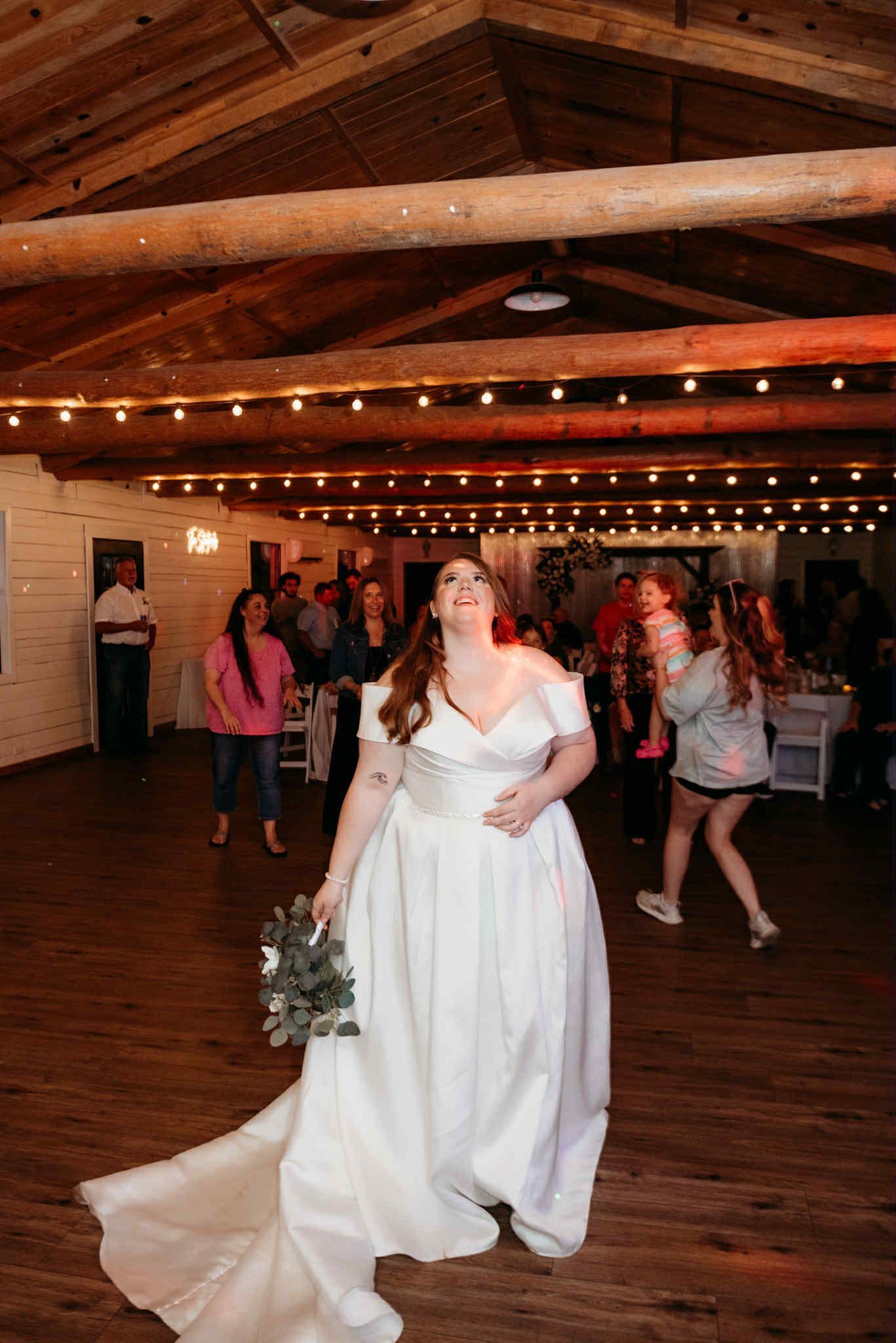 Bride tossing bouquet at a wedding reception. White dress, wooden interior, fairy lights.