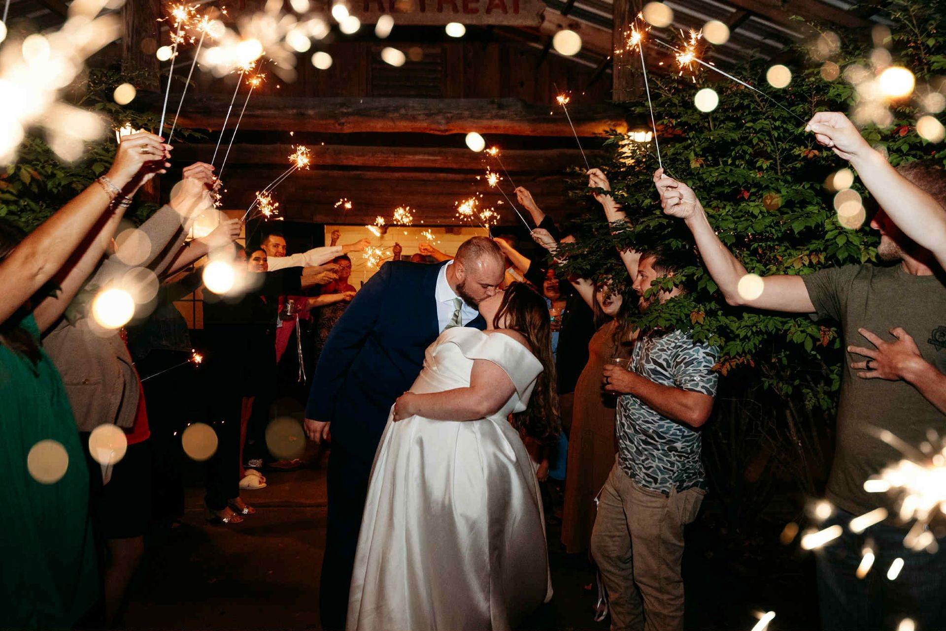 Couple kissing under sparklers, wedding send-off. Guests hold sparklers in an outdoor venue.