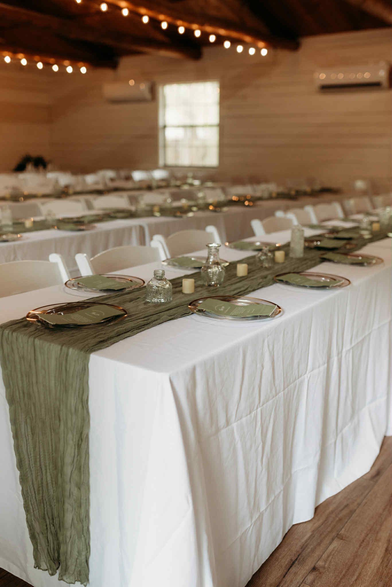 Long tables set for a gathering with white tablecloths, olive green runners, and place settings inside a bright, rustic room.