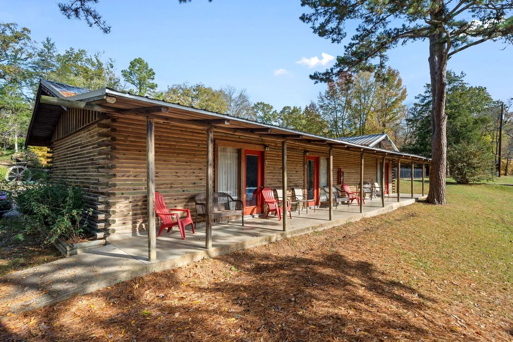 Log cabin with red doors and chairs on a porch, surrounded by trees and grass.