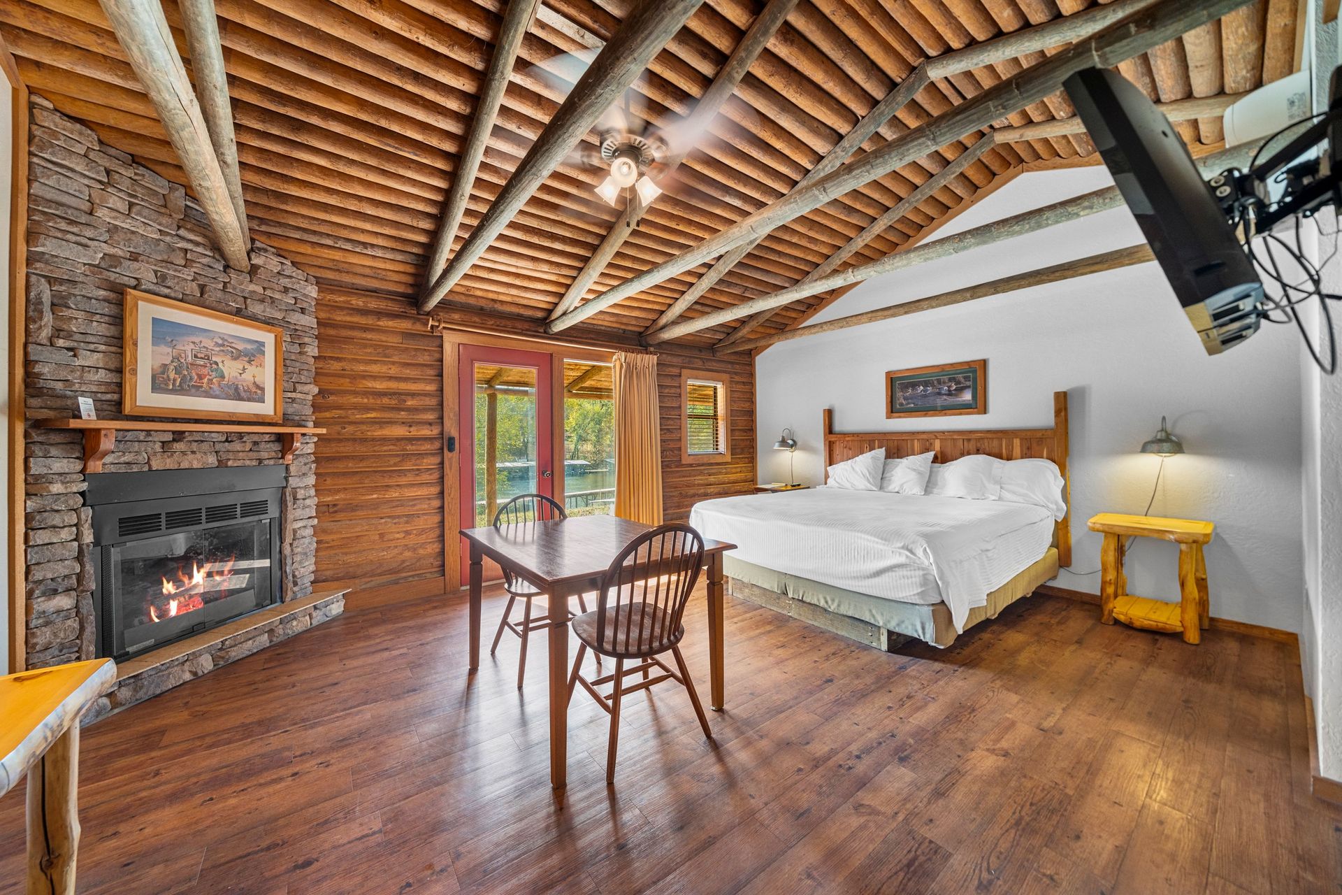 Rustic lodge bedroom with fireplace, bed, and wooden table. Exposed beams, wood floors, and a TV.
