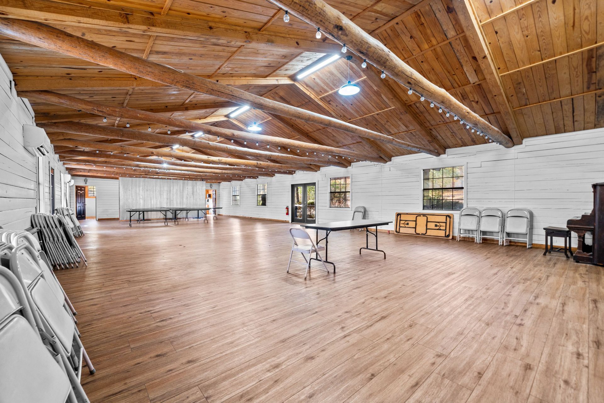 Wide interior of a wooden-floored hall with white walls, exposed beams, and folding chairs.