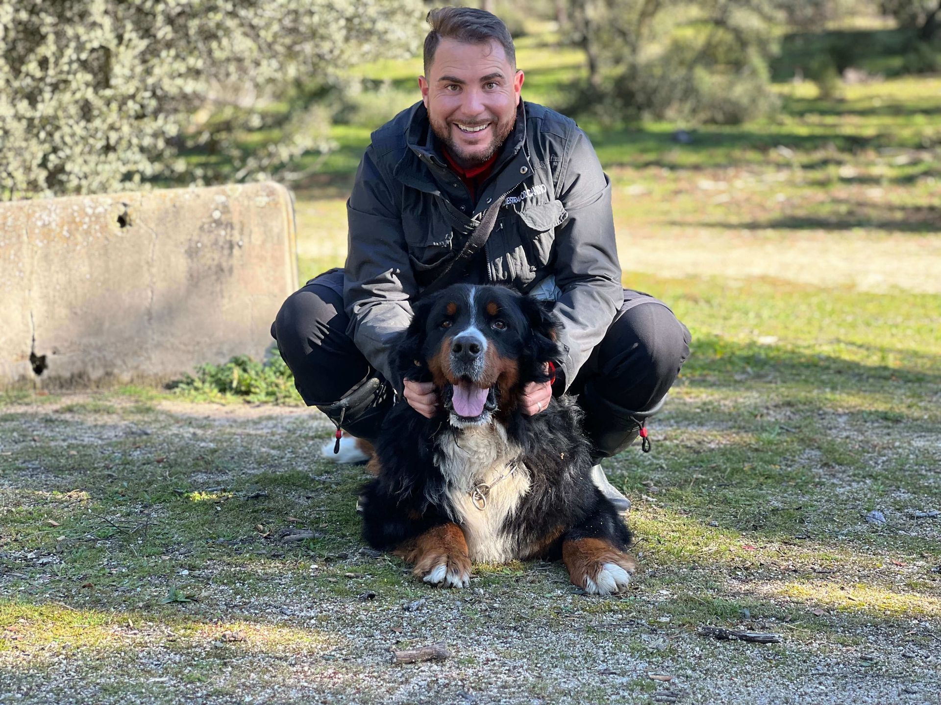 Una persona con una chaqueta oscura está en cuclillas en una zona de césped al aire libre, sonriendo mientras sostiene un gran perro de montaña bernés.