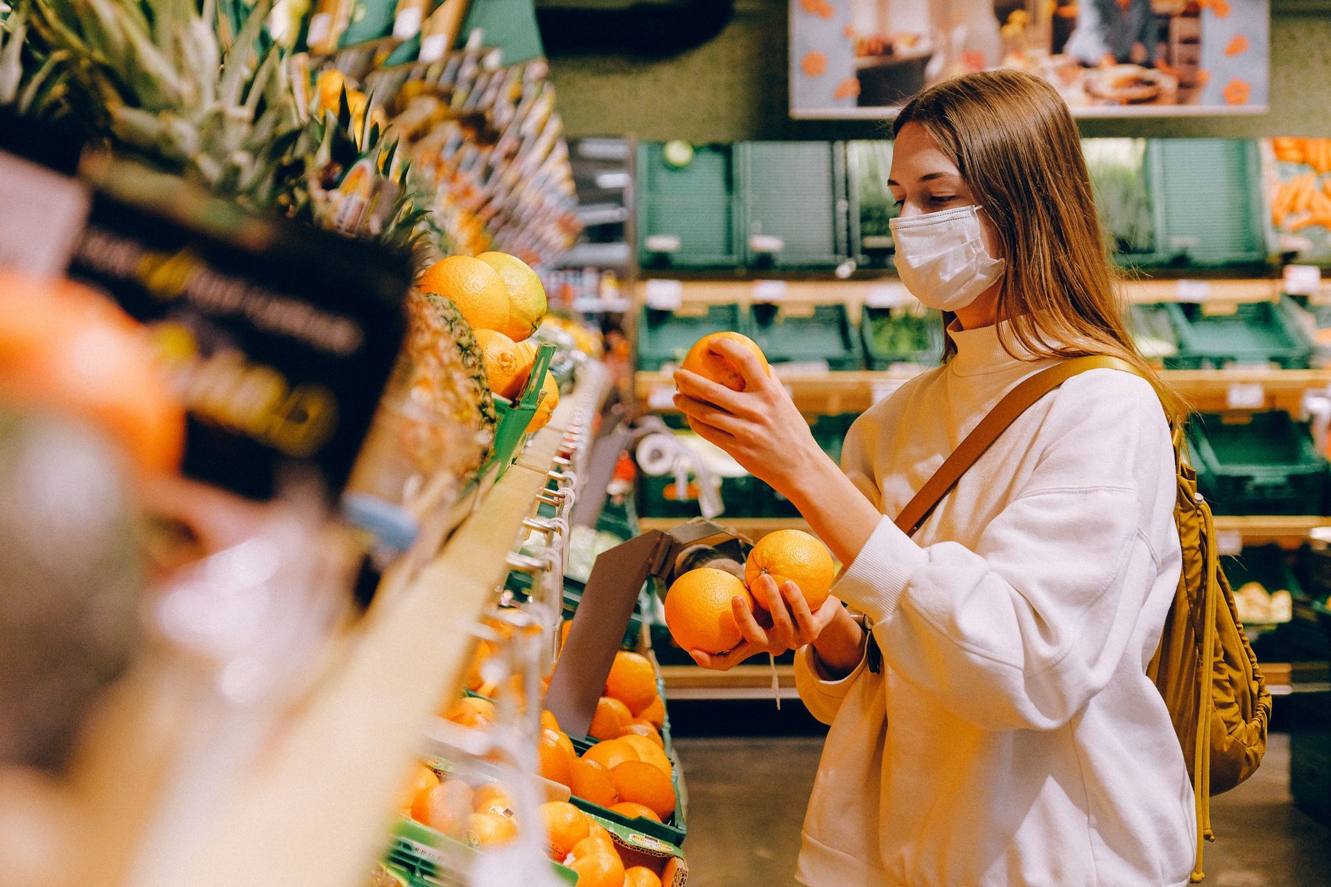 A woman wearing a face mask is buying oranges in a grocery store.