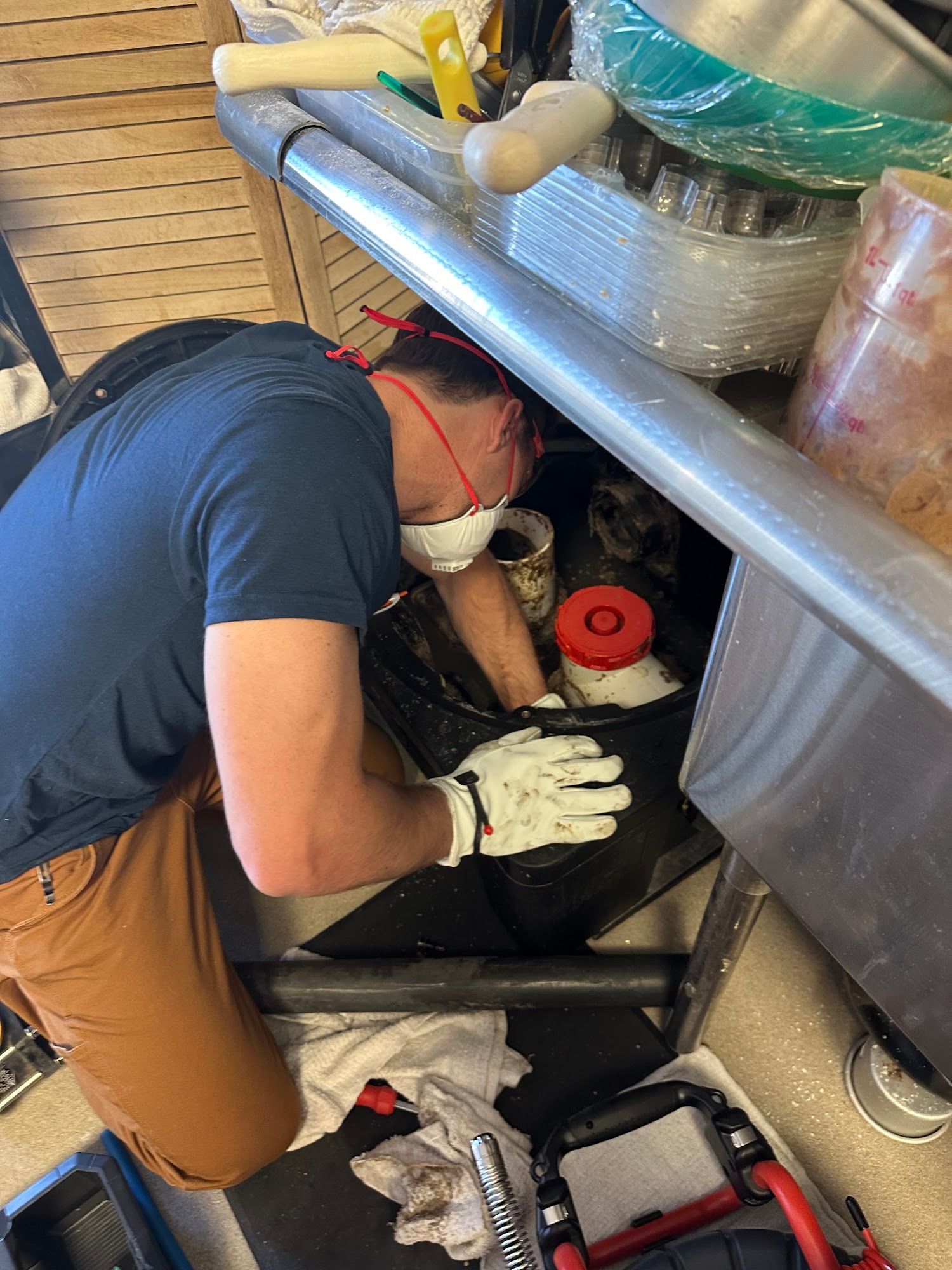 Man in mask and gloves under sink, reaching into a trash bin with containers.