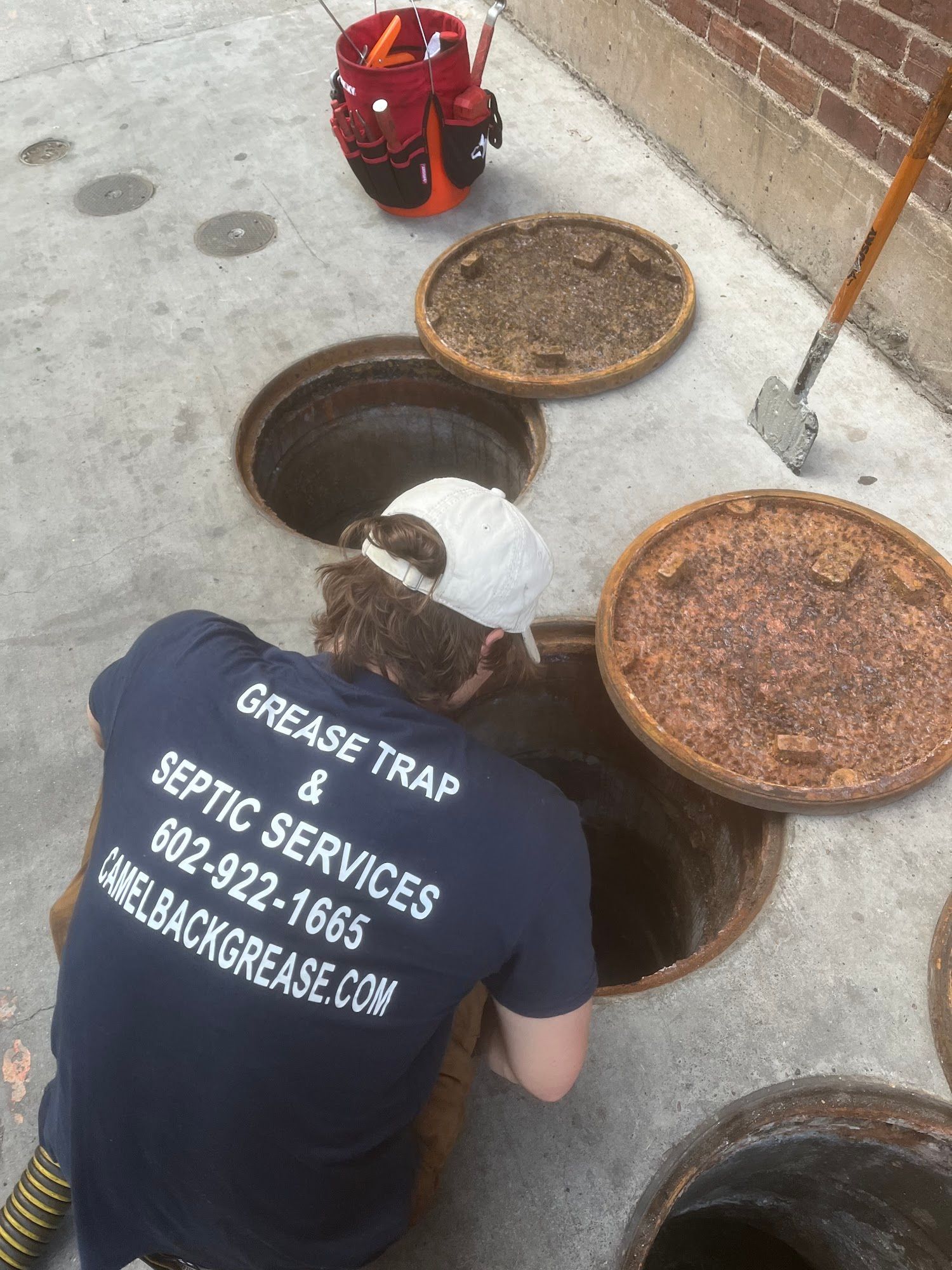 Man cleaning grease trap, Camelback Grease Services. Open manholes, tools nearby, brick wall.