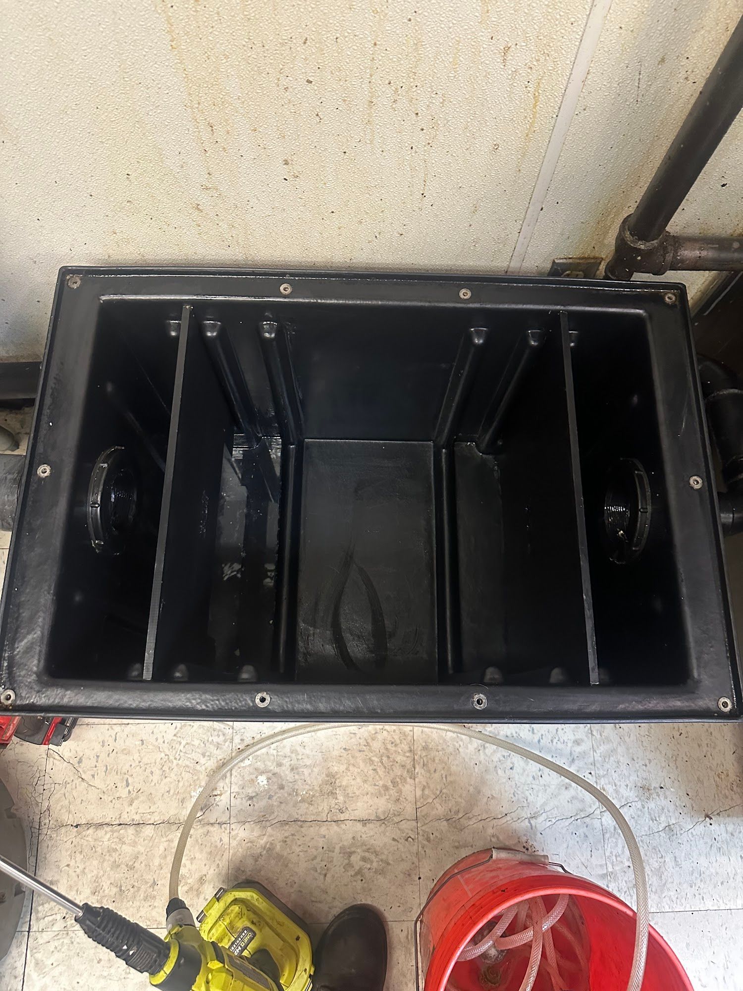 Black rectangular tank with internal dividers, near a hose, bucket, and power tool.