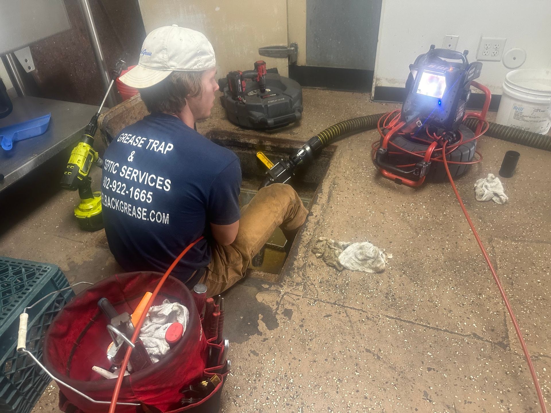 Plumber in a utility room, inspecting a drain with a camera. Tools in red bucket; black drain cleaning equipment.