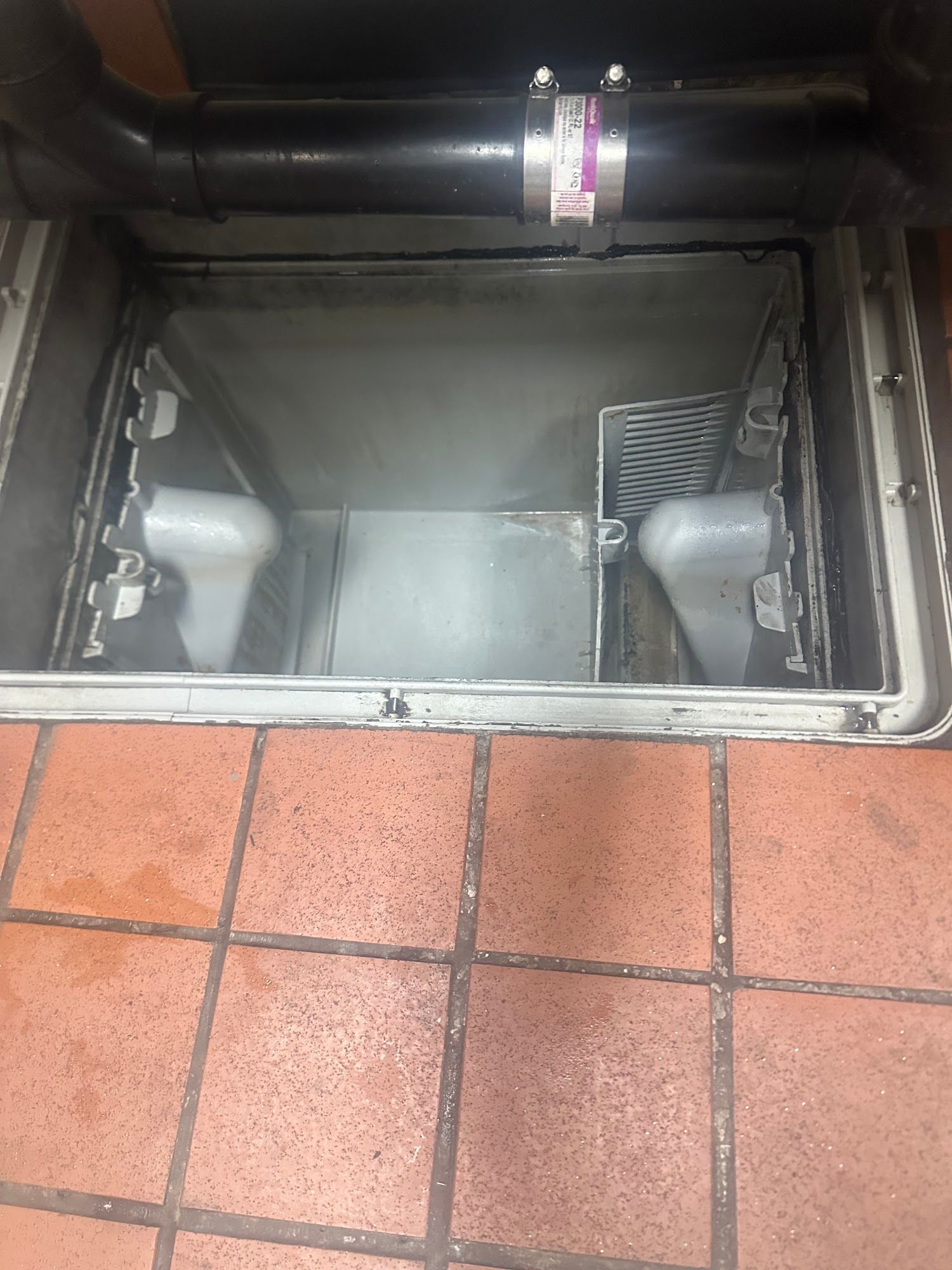 A rectangular floor drain with metal components. Black pipe overhead, set on red tiled floor.