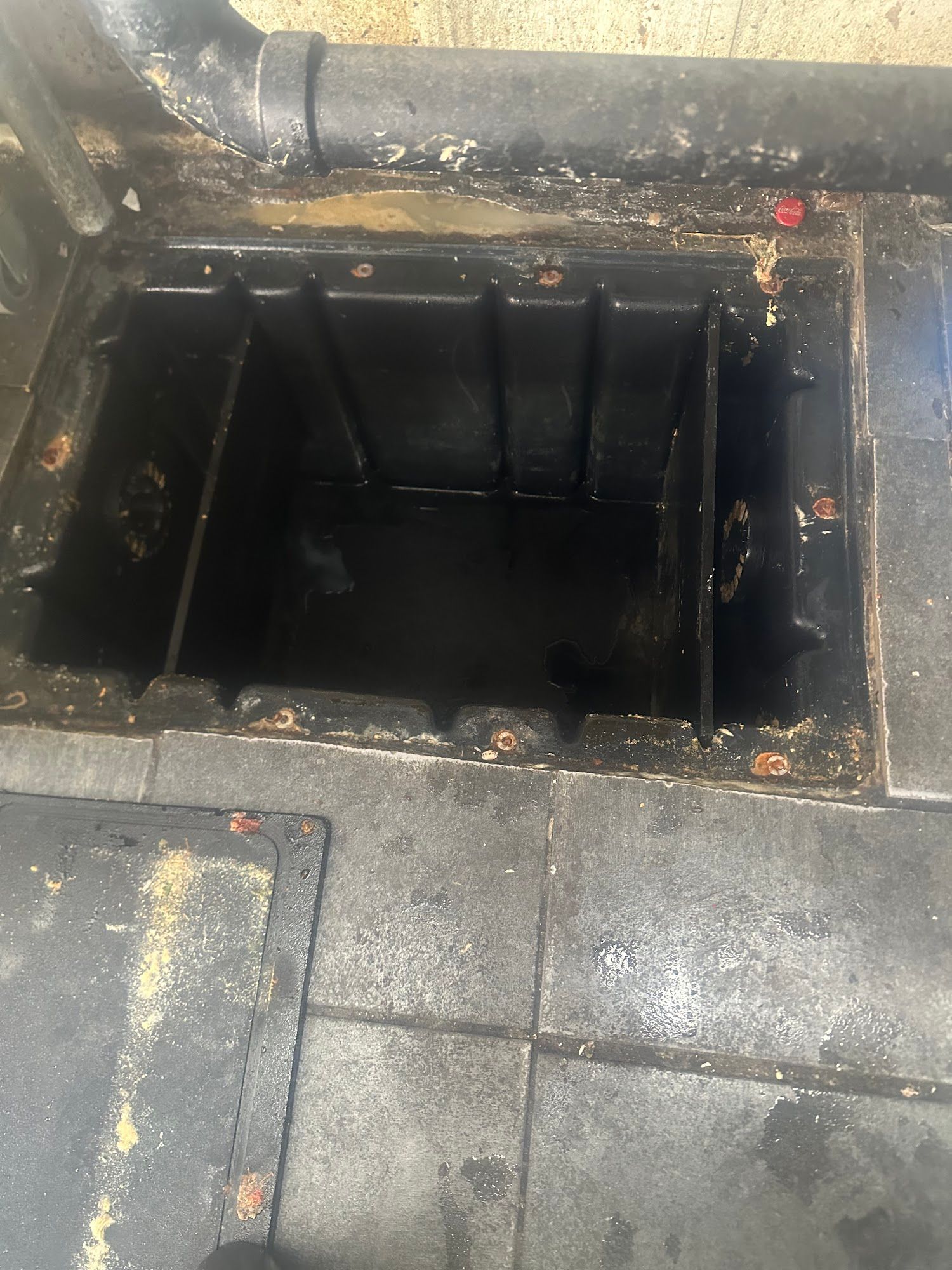 Black rectangular drain with interior compartments, set in dark, tiled flooring. A pipe extends from above.
