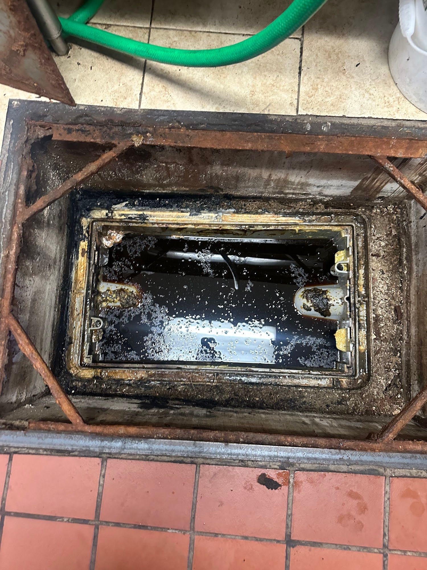 Open grease trap in a commercial kitchen floor, filled with dark oil and debris.