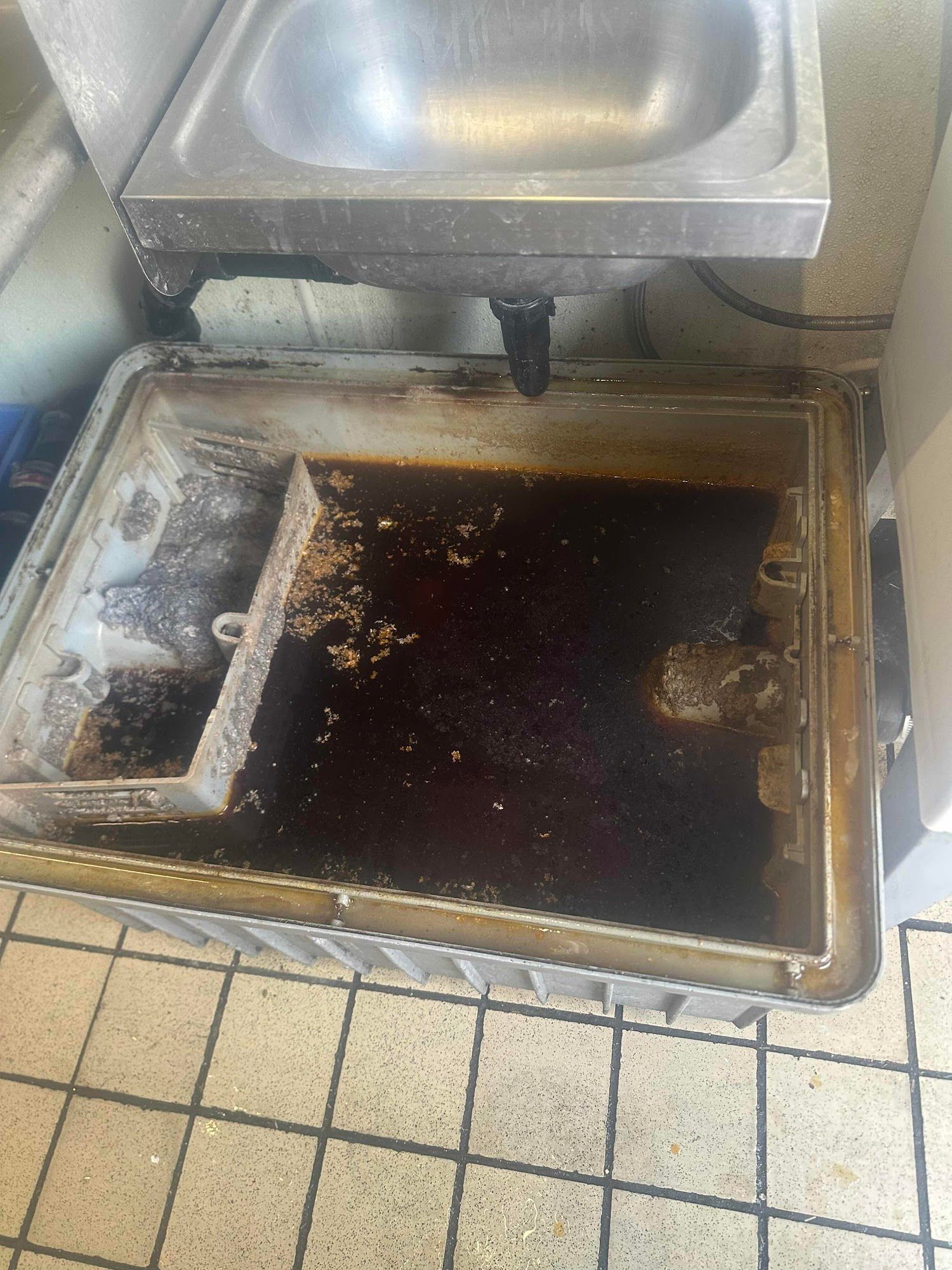 Dirty, greasy catch basin under a commercial sink. The basin is filled with dark, oily liquid.