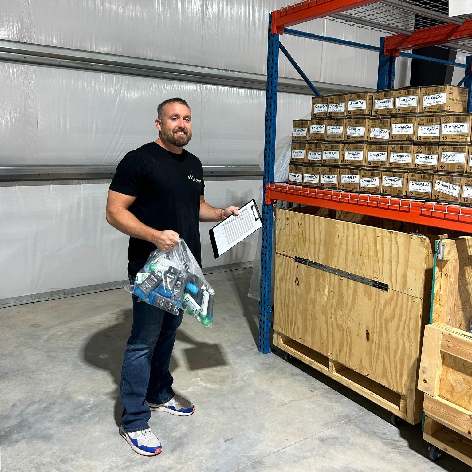 A man is standing in a warehouse holding a clipboard and a bag of bottles.