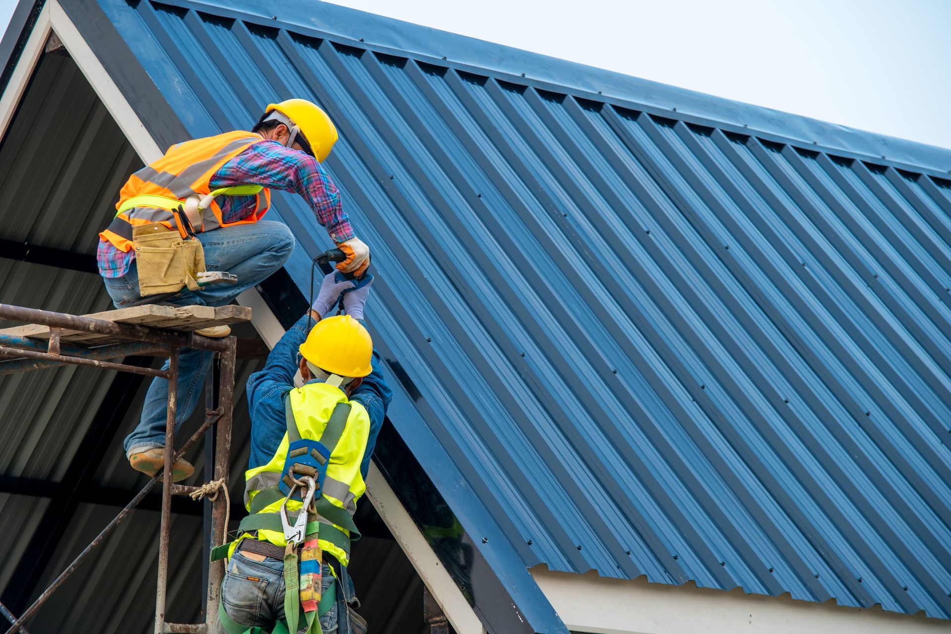 Two construction workers are working on a blue metal roof.