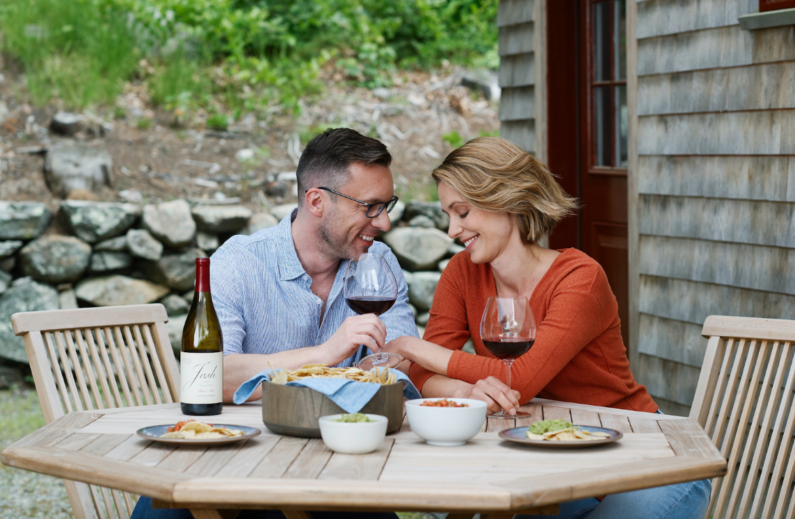 Couple enjoying wine and a meal outdoors at a wooden table. 