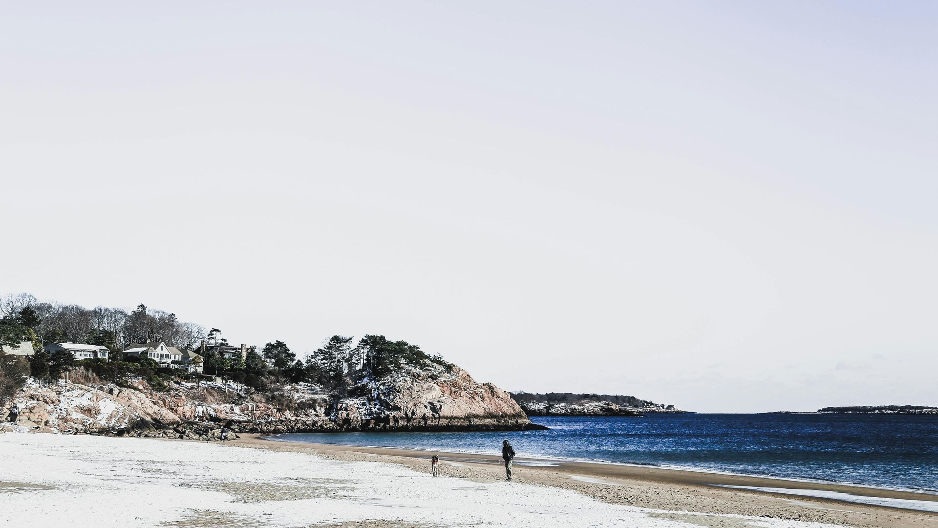 Beach scene with blue water, sandy shore, rocky cliff with trees, and person walking.