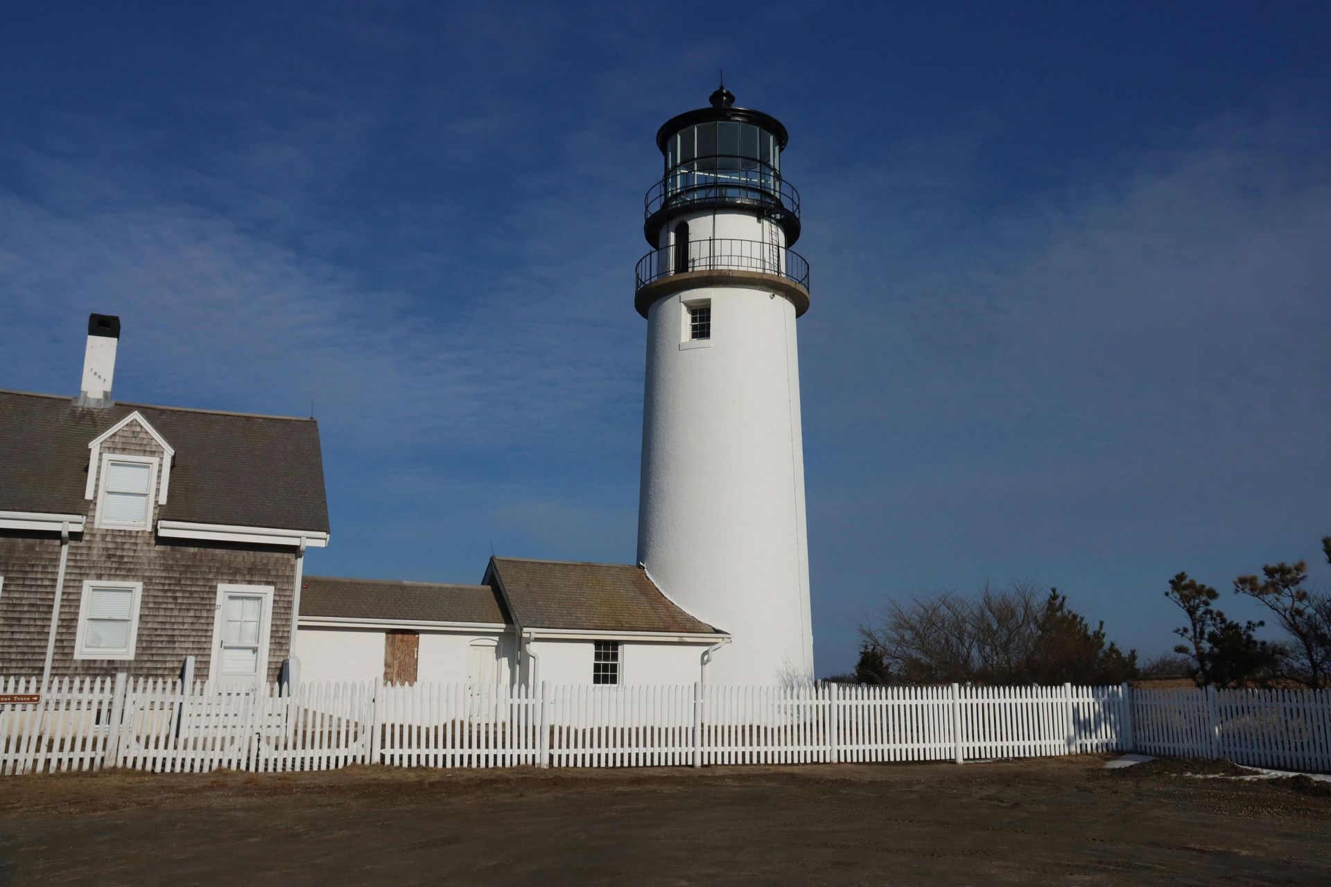 White lighthouse next to a small wooden building and picket fence on a sunny day.