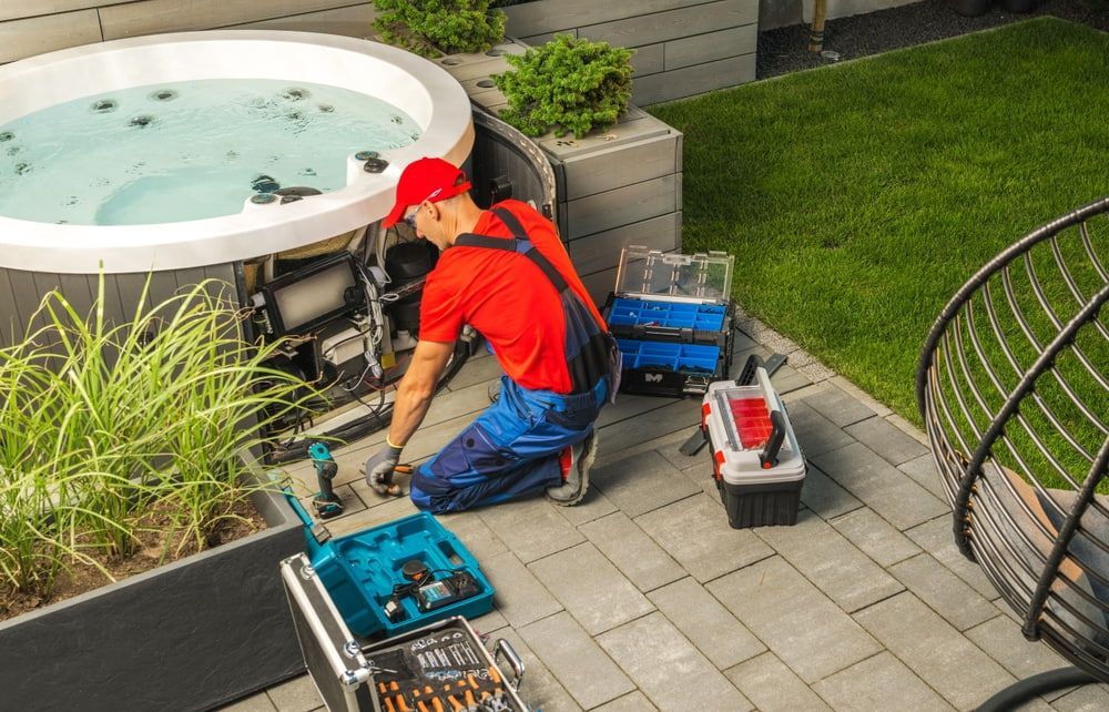 A person in red and blue work clothes repairs a hot tub outdoors, with tools nearby