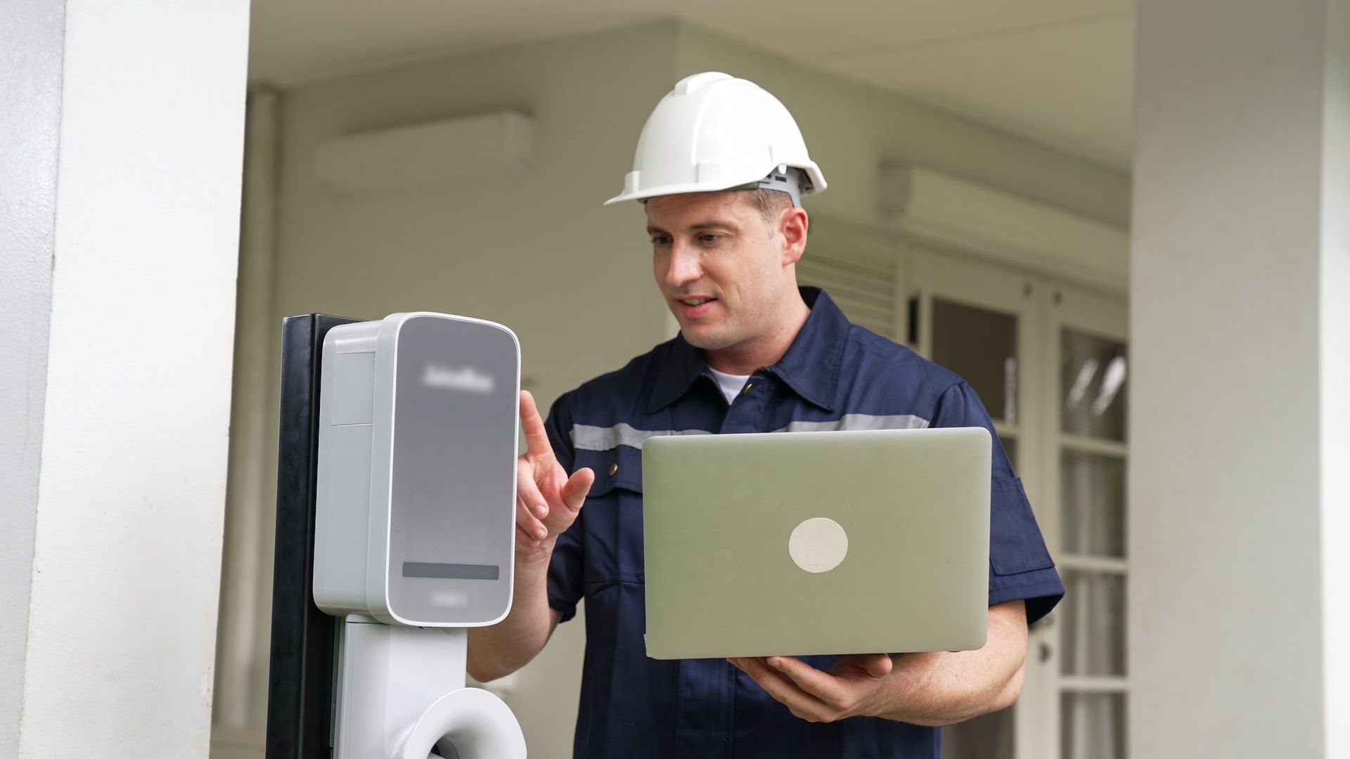 Man in hard hat and work uniform interacts with a device, holding a laptop outside a building