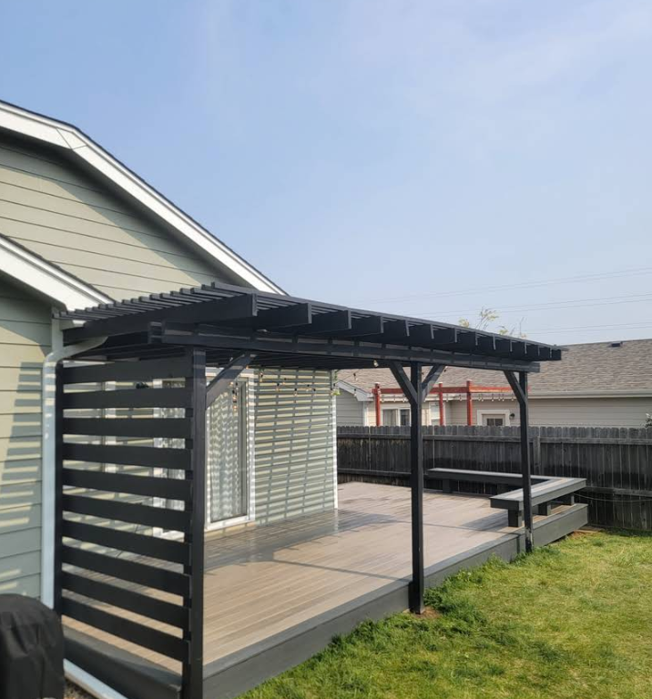 A black wooden pergola and slatted privacy screen on a backyard deck attached to a light green house.