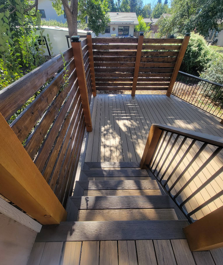 A high-angle view looking down a wooden staircase onto a deck with a horizontal-slat wooden privacy screen.