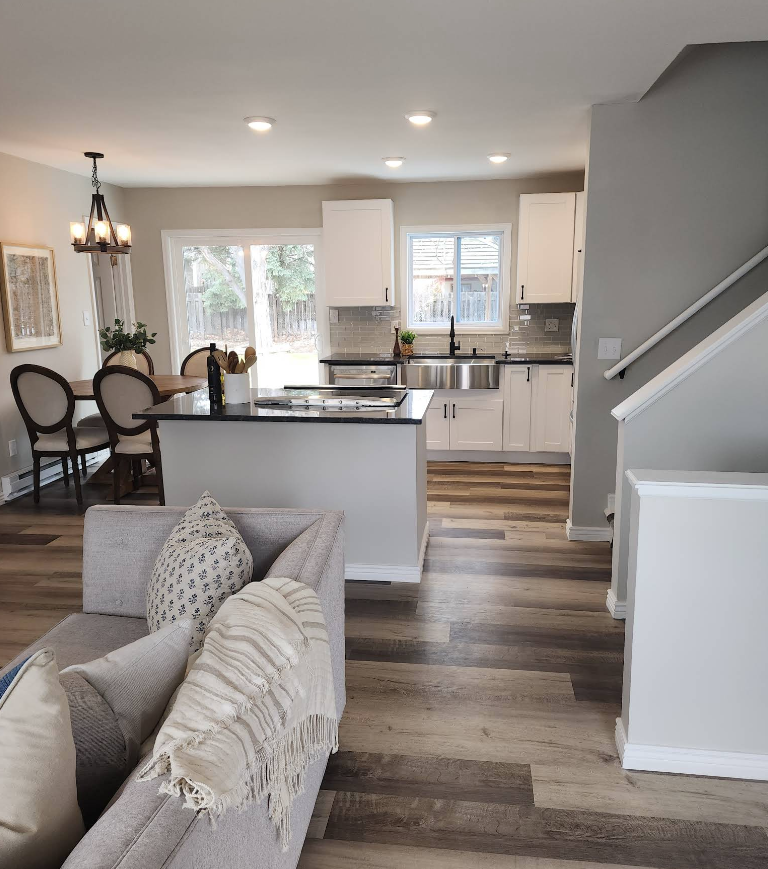 A view from a living room toward a kitchen with white cabinets, a black island, dining area, and wood-look flooring.