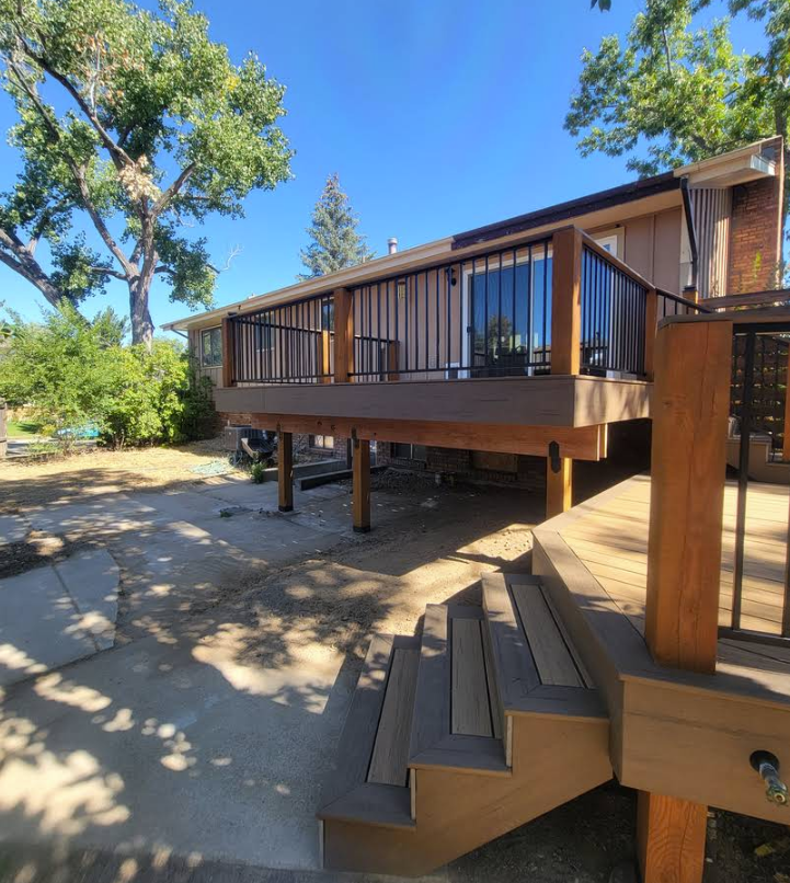 An elevated wooden deck with a black metal railing stands over a concrete patio, featuring matching stairs in the foreground.