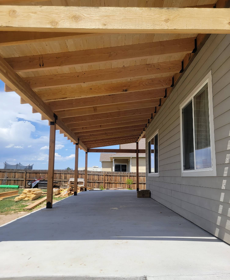 A newly built covered concrete patio attached to a gray-sided house, featuring wooden beams and support posts.