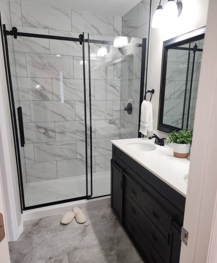 Modern bathroom with a black-framed glass shower, marble-tiled walls, a dark wood vanity, and a potted plant.