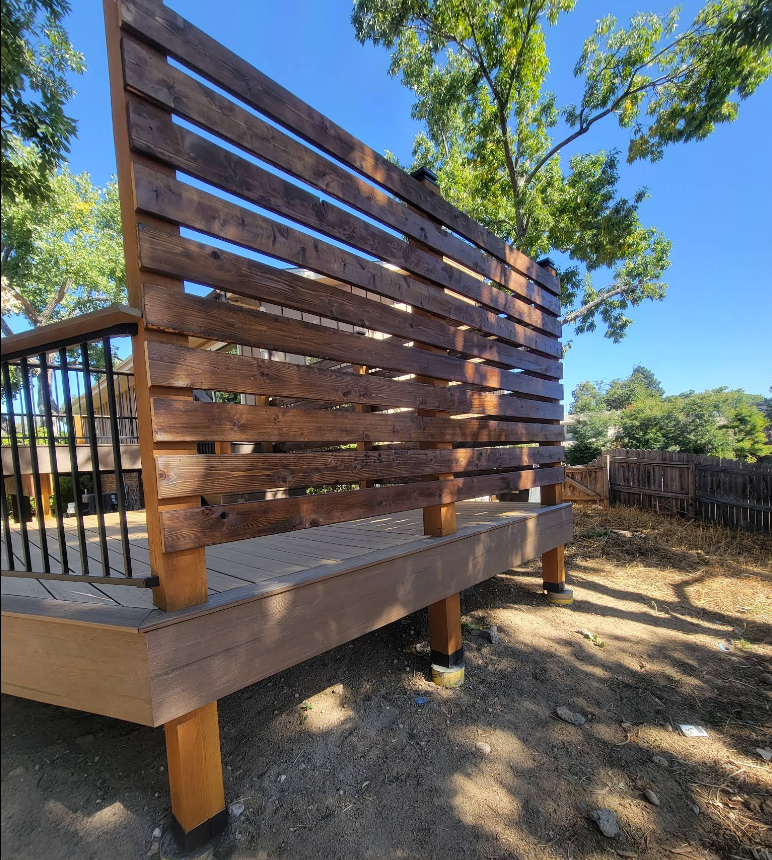 A stained wooden privacy screen with horizontal slats on a backyard deck, viewed from below in a sunny outdoor setting.