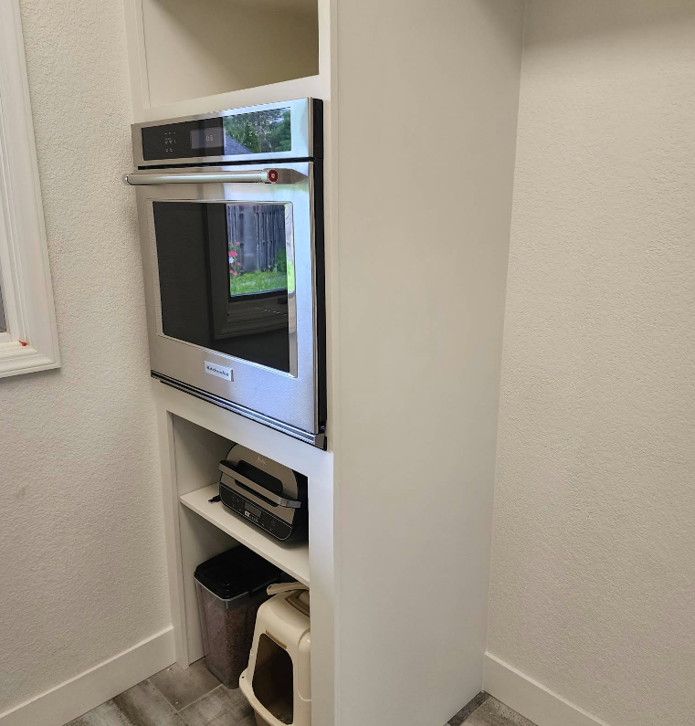 A built-in kitchen cabinet featuring an oven, an open shelf holding a toaster, and a bottom section with a pet carrier.