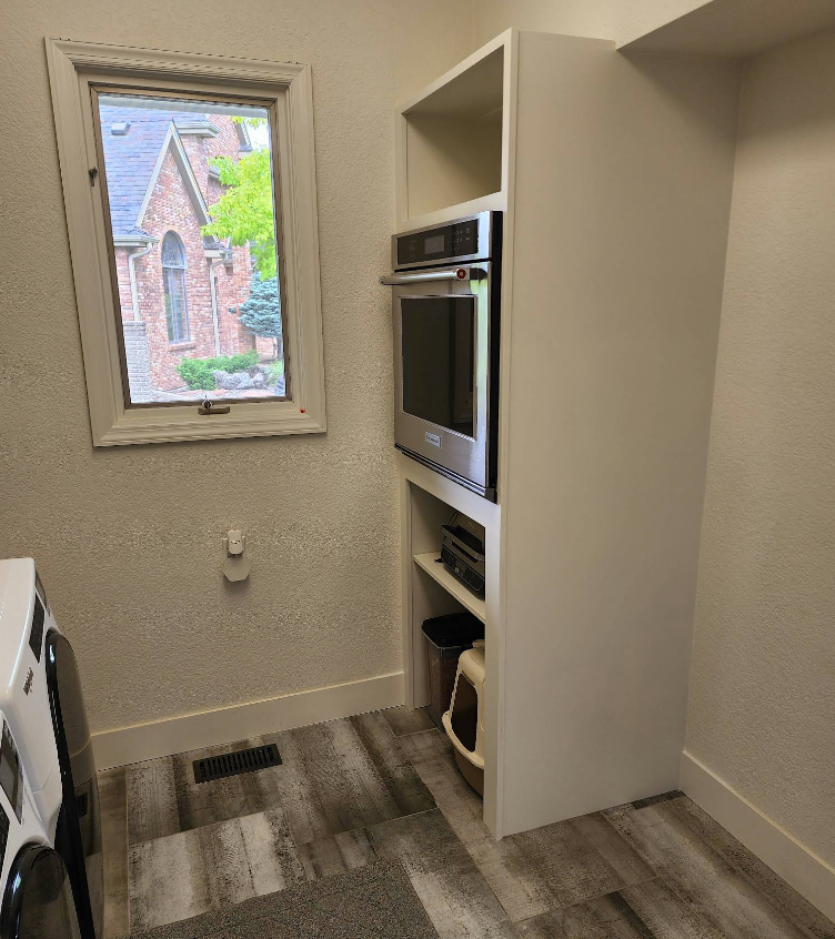A laundry room with a washing machine next to a built-in white cabinet housing an oven, with a window on the left.