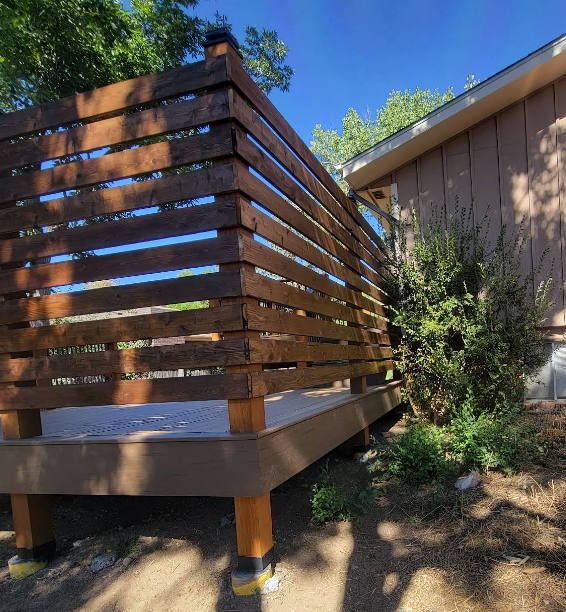 A wooden deck with a horizontal-slat privacy screen, raised on posts near a house wall and a small green bush.