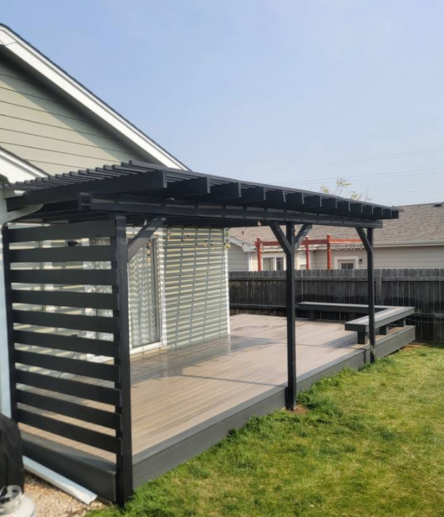 A black wooden pergola and slatted privacy screen on a gray deck attached to the side of a house with a grass yard.