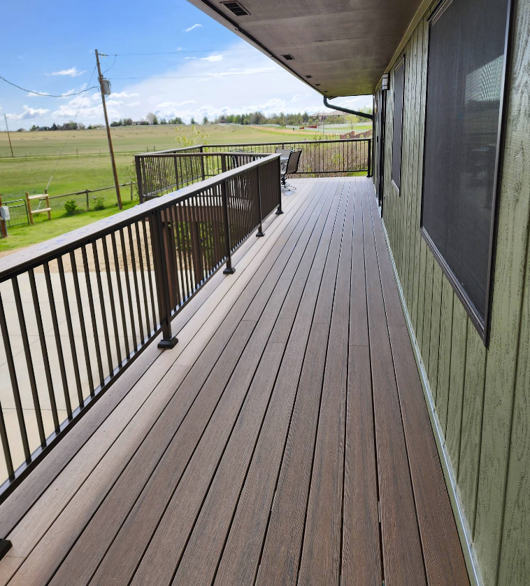 A long wooden deck with black railings overlooks a grassy field under a bright, sunny sky.