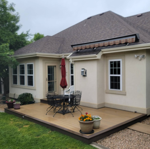 A beige stucco house with a dark brown deck, a patio table with chairs, an umbrella, and potted plants in the backyard.