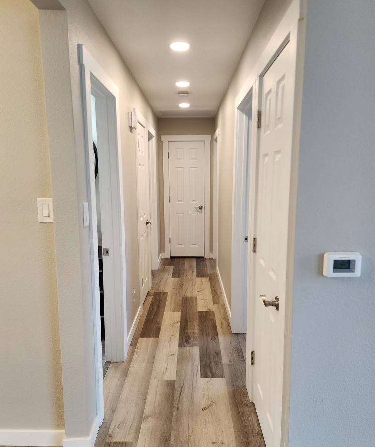 A hallway with light-colored walls and wood-patterned flooring leads to a white door, with three recessed ceiling lights.