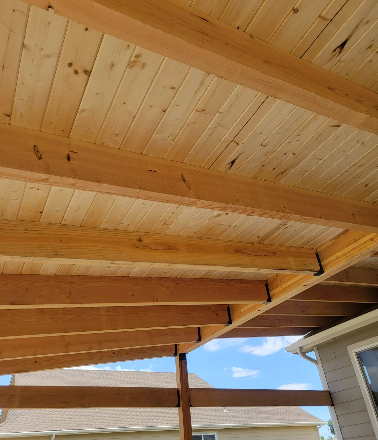 A view looking up at a wooden ceiling structure with exposed beams and tongue-and-groove boards on a patio.