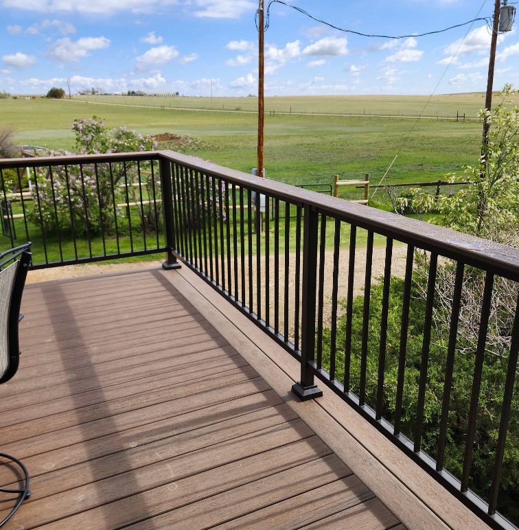 A brown composite deck with a black metal railing overlooking a vast, grassy rural landscape under a blue sky.