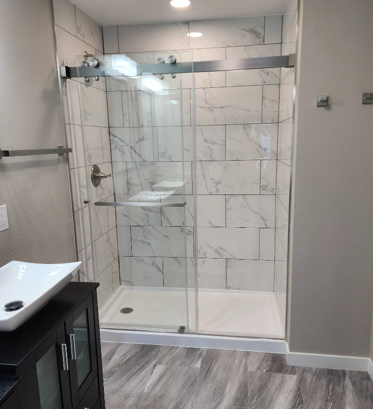 A modern bathroom with a glass sliding shower door, white marble-patterned tile walls, a shower pan, and a vanity cabinet.