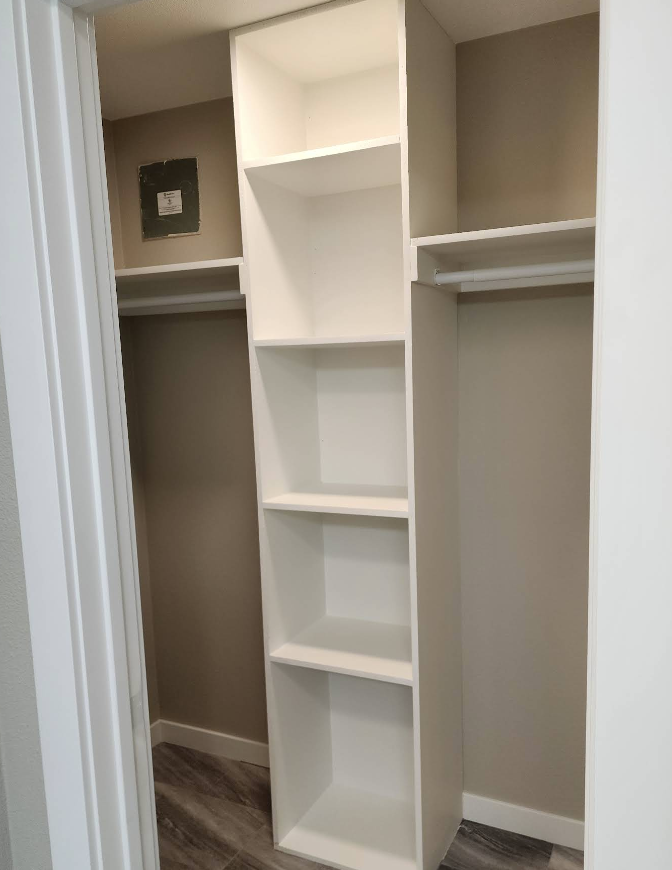 A white built-in shelving unit centered between two closet spaces with hanging rods on beige walls and wood-look flooring.