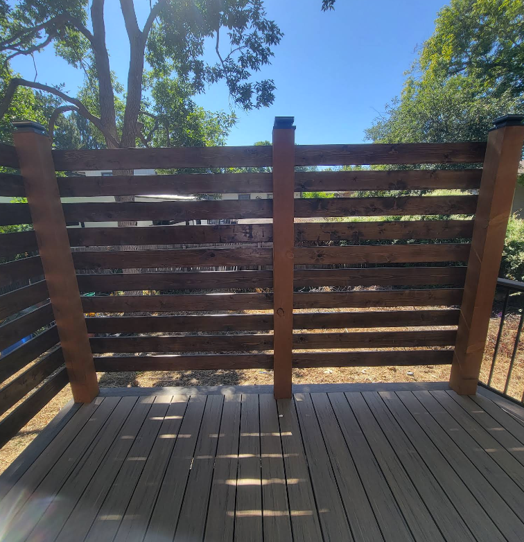 A wooden privacy screen with horizontal slats stands on a grey deck against a background of trees and blue sky.