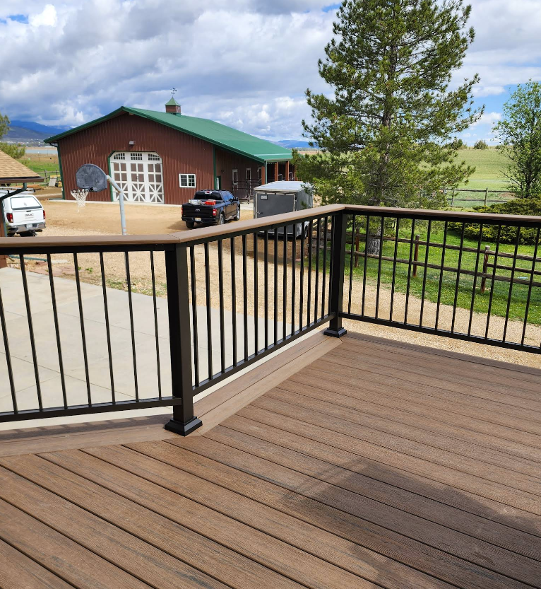 A brown composite deck with a black railing overlooks a yard with a red barn, a parked truck, and trees under a cloudy sky.