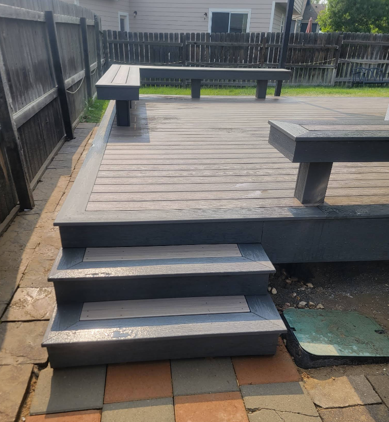 A gray wooden deck with built-in bench seating and two wide steps leading down to a paved patio in a fenced backyard.
