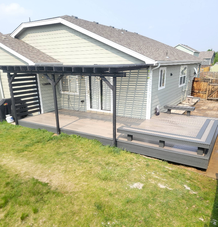 A light green house with a dark gray patio deck, built-in benches, and a black wooden pergola over the sliding doors.
